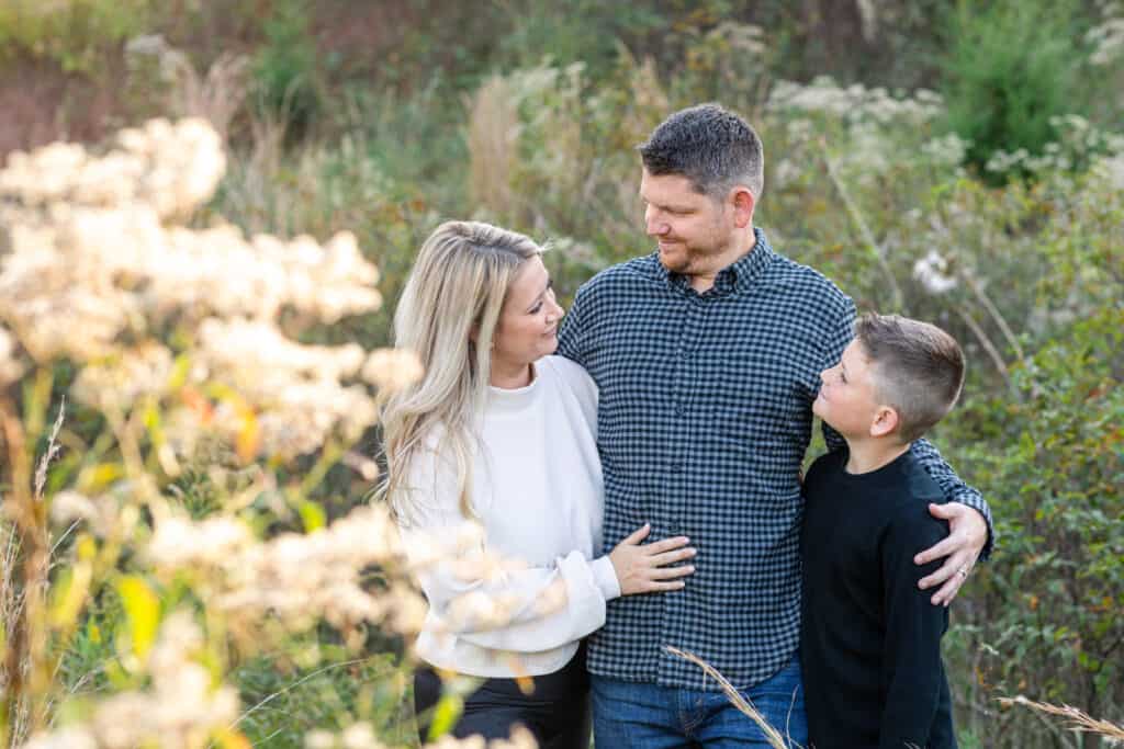 Mother, Father and son stand and look at each other and smile in grassy field.